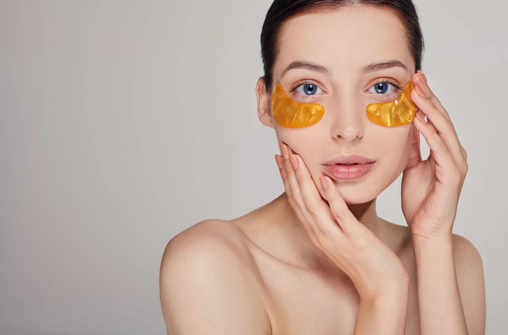 Pritty girl on the background applying golden collagen patches under the eyes. Mask for removing wrinkles and dark circles. A woman takes care of delicate skin around her eyes. Cosmetic procedures. - Photo, Image