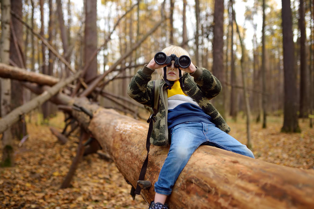 Escoteiro com binóculos durante caminhadas na floresta de outono. Criança está sentada em uma grande árvore caída e olhando através de um binóculos
. - Foto, Imagem