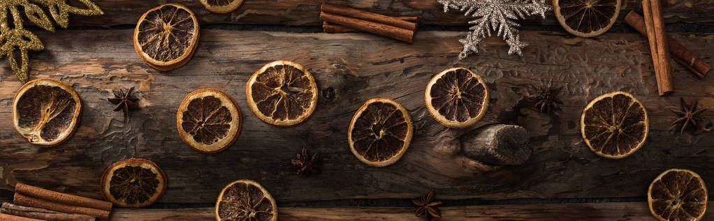 vrchní pohled na sušené citrusové plátky s anýzem, skořicové tyčinky a dekorativní sněhové vločky na dřevěném pozadí, panoramatický záběr - Fotografie, Obrázek
