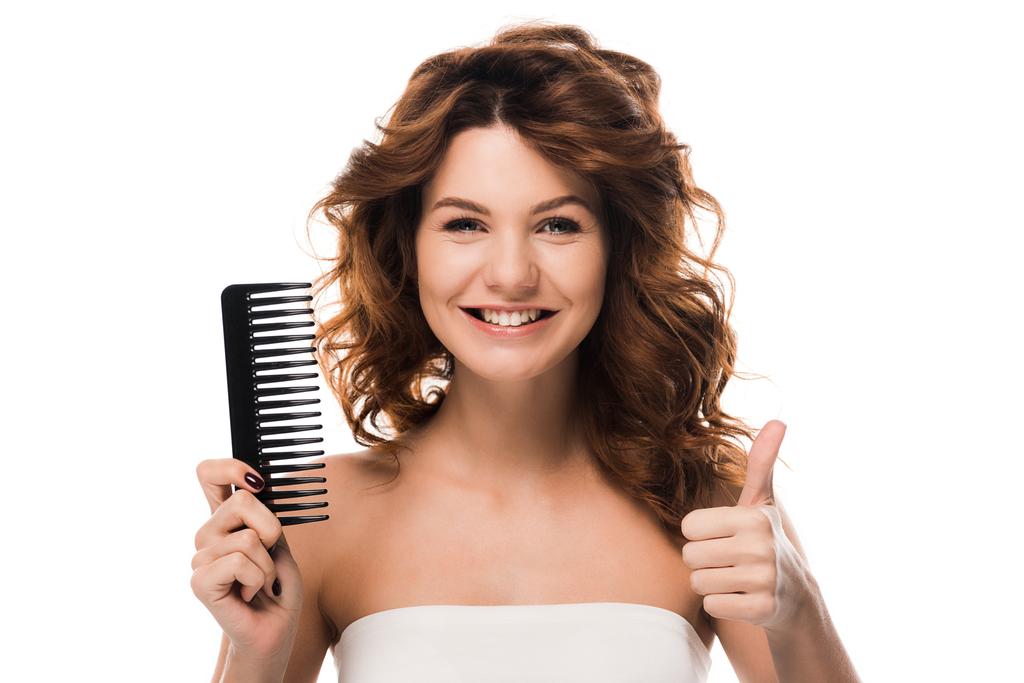 glückliches Mädchen mit lockigem Haar hält Haarbürste und zeigt Daumen nach oben isoliert auf weiß  - Foto, Bild