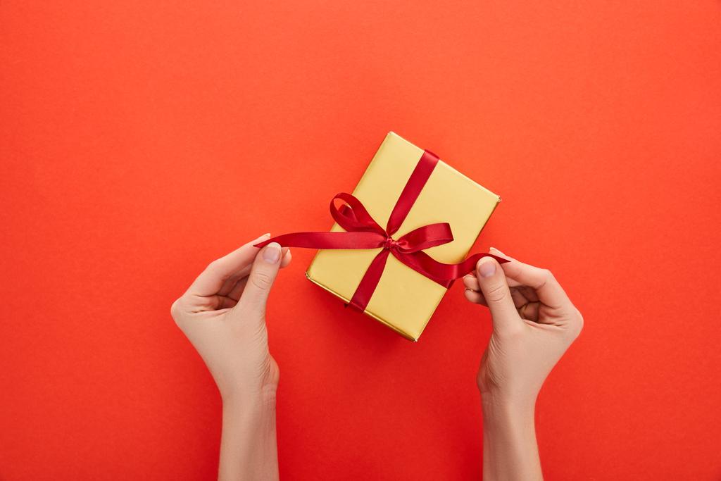 vista recortada de la mujer desatar cinta en caja de regalo de oro sobre fondo rojo
 - Foto, imagen