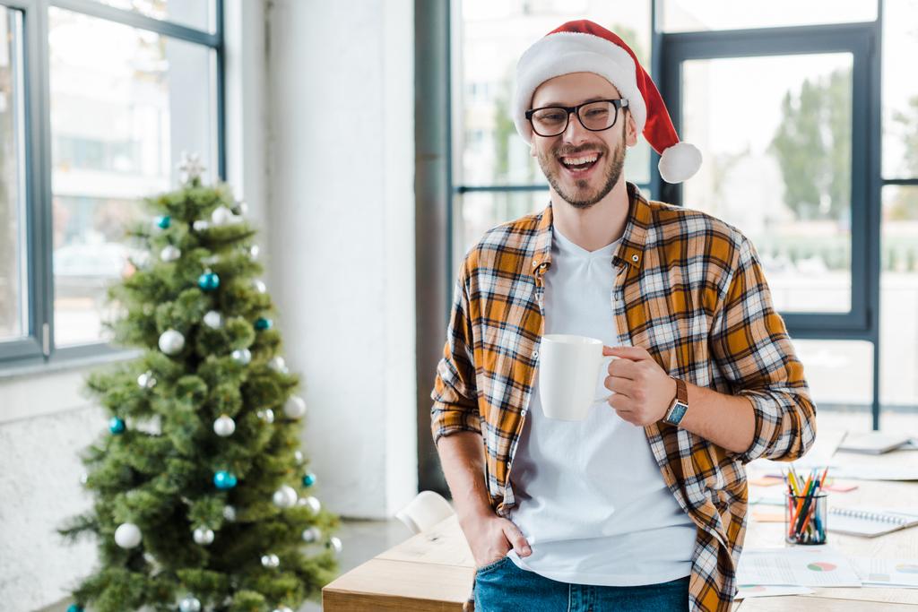 selective focus of cheerful bearded man in santa hat holding cup near christmas tree in office  - Photo, Image