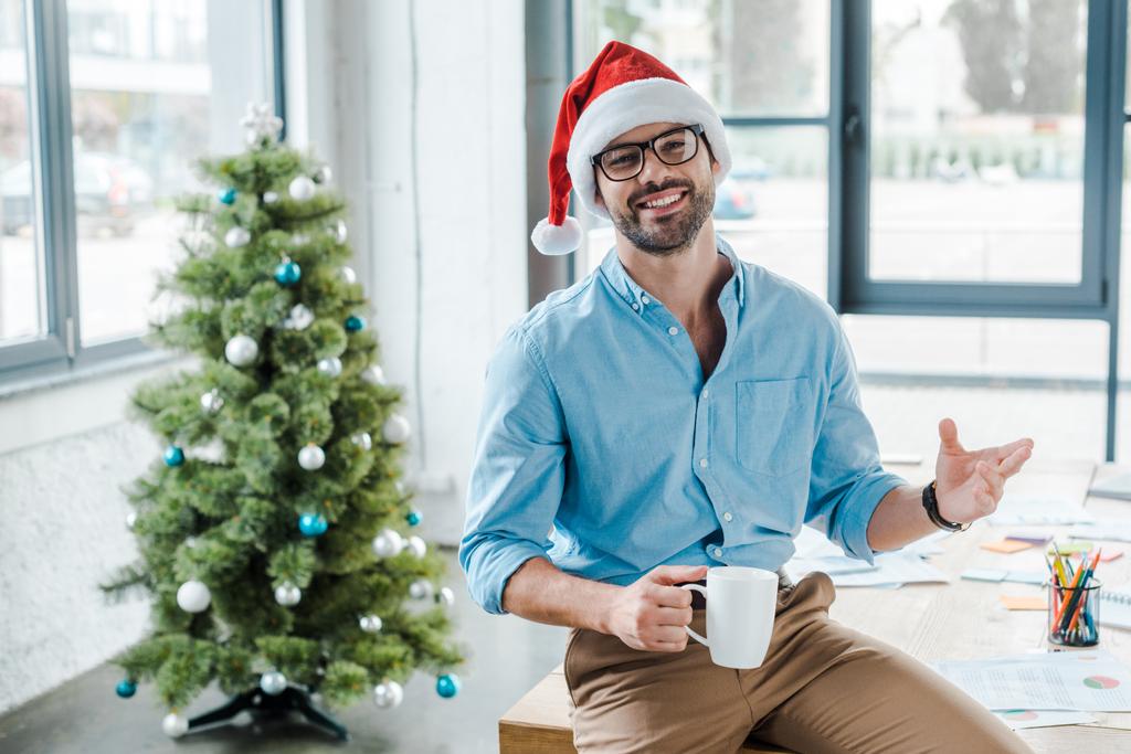 enfoque selectivo de hombre barbudo alegre en sombrero de santa y vasos sosteniendo taza con café cerca del árbol de Navidad en la oficina
  - Foto, imagen