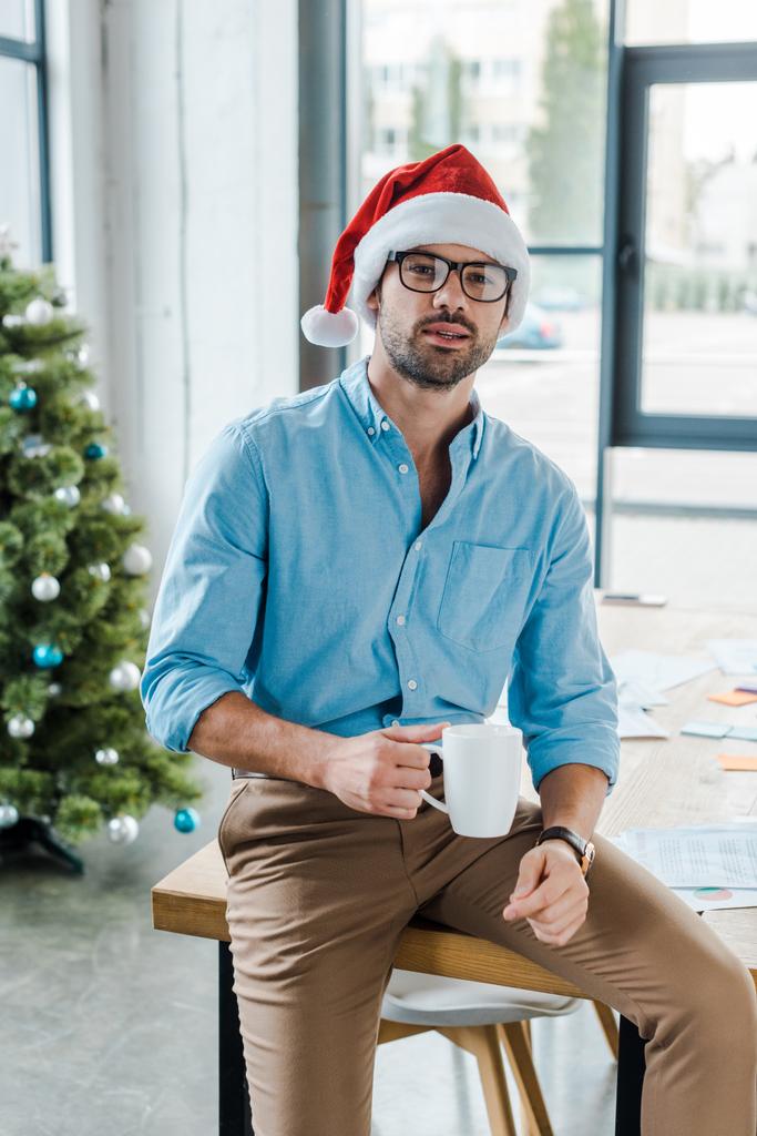 επιλεκτική εστίαση του γενειοφόρου άνδρα σε καπέλο Σάντα και ποτήρια κρατώντας κύπελλο με καφέ κοντά στο χριστουγεννιάτικο δέντρο στο γραφείο  - Φωτογραφία, εικόνα