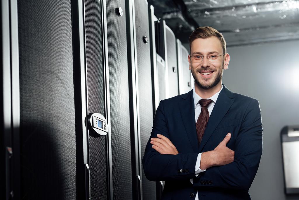 selective focus of cheerful businessman in suit and glasses standing with crossed arms in data center  - Photo, Image