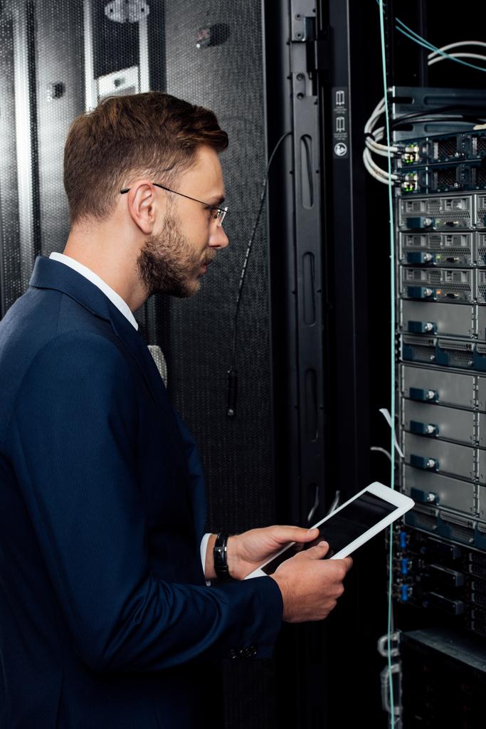 handsome businessman in glasses holding digital tablet and looking at database in server rack  - Photo, Image