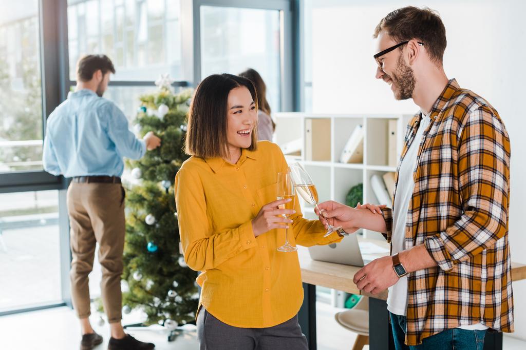 selective focus of happy businessman and asian businesswoman toasting champagne glasses near colleagues and christmas tree  - Photo, Image