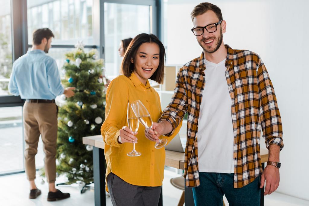 Selektiver Fokus des fröhlichen Geschäftsmannes und der asiatischen Geschäftsfrau, die Champagnergläser neben Kollegen und Weihnachtsbaum anstoßen  - Foto, Bild