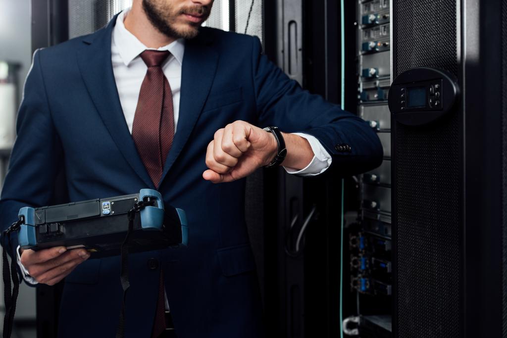 обрезанный вид бизнесмена стоящего с рефлектометром и смотрящего на часы в серверной
  - Фото, изображение