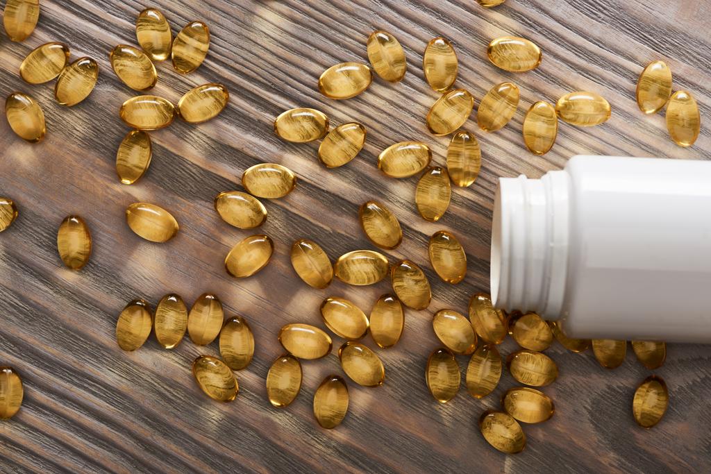 cápsulas de óleo de peixe dourado espalhadas do recipiente de plástico na mesa de madeira
 - Foto, Imagem