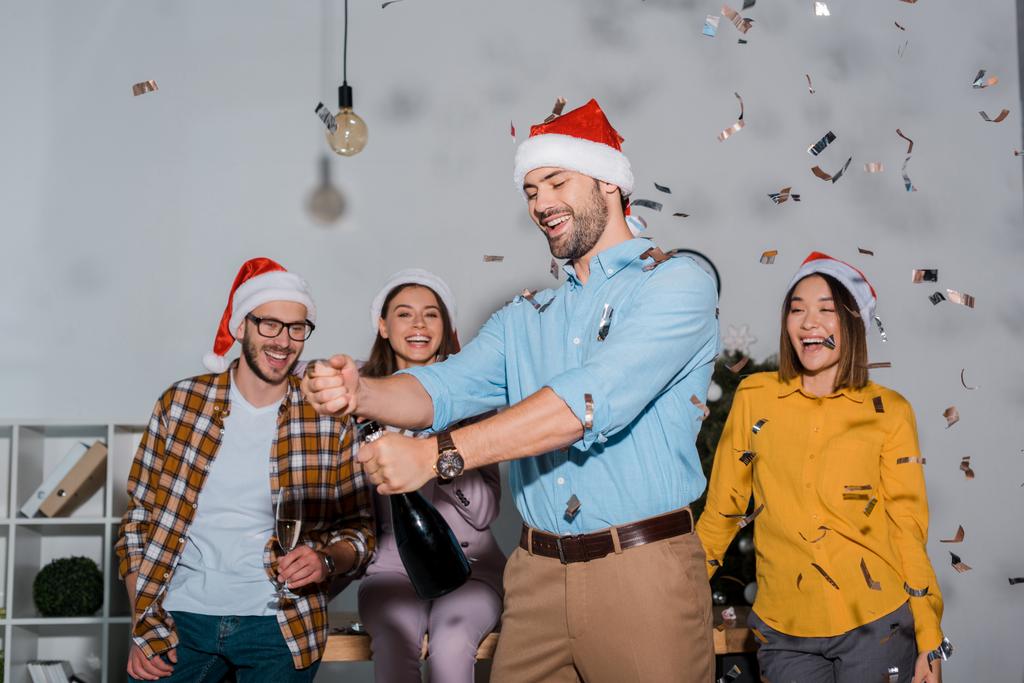 szczęśliwy biznesmen w kapeluszu Santa trzymając butelkę szampana w pobliżu współpracowników i spadające confetti  - Zdjęcie, obraz