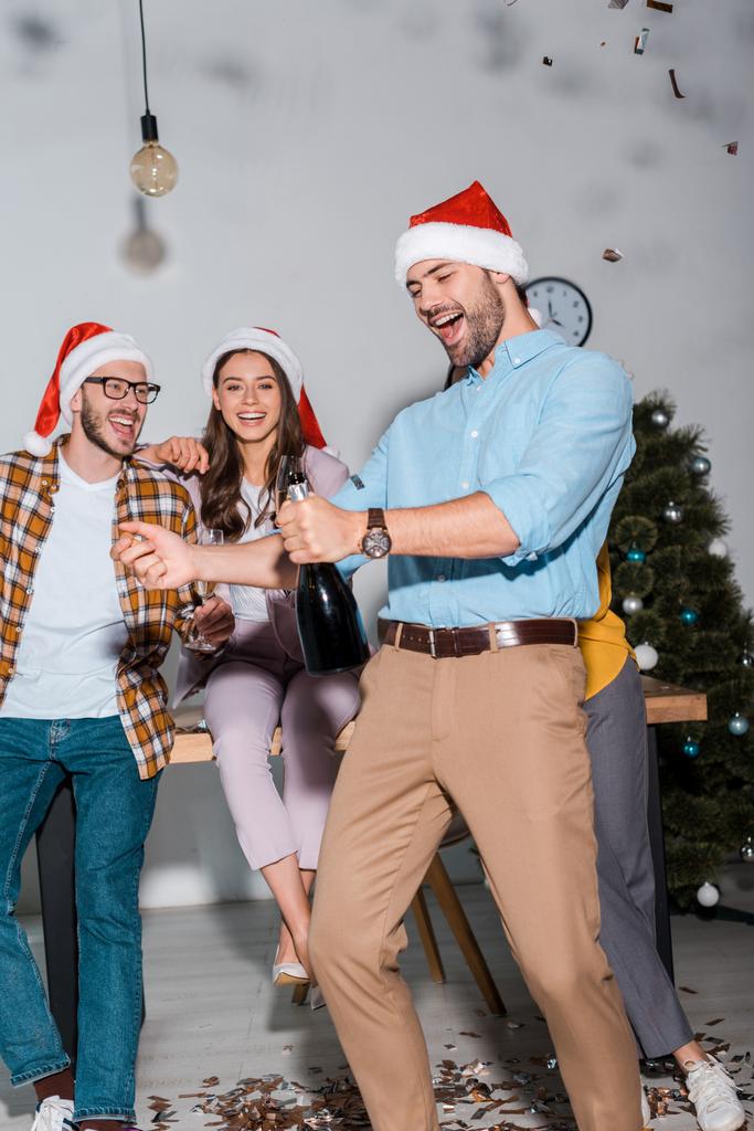 szczęśliwy biznesmen w kapeluszu Santa trzymając butelkę szampana w pobliżu współpracowników i spadające confetti  - Zdjęcie, obraz