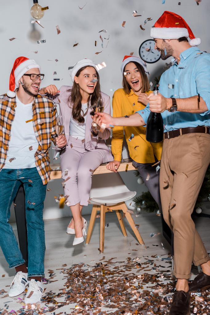 happy businessmen and multicultural businesswomen in santa hats with champagne glasses near falling confetti  - 写真・画像