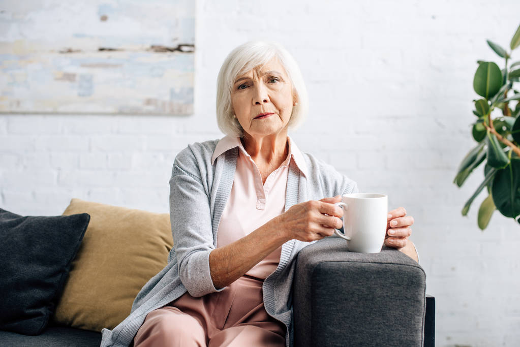 ηλικιωμένη γυναίκα κάθεται στον καναπέ και κρατώντας κύπελλο στο διαμέρισμα  - Φωτογραφία, εικόνα
