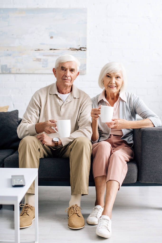 smiling husband and wife holding cups and sitting on sofa in apartment  - Photo, Image
