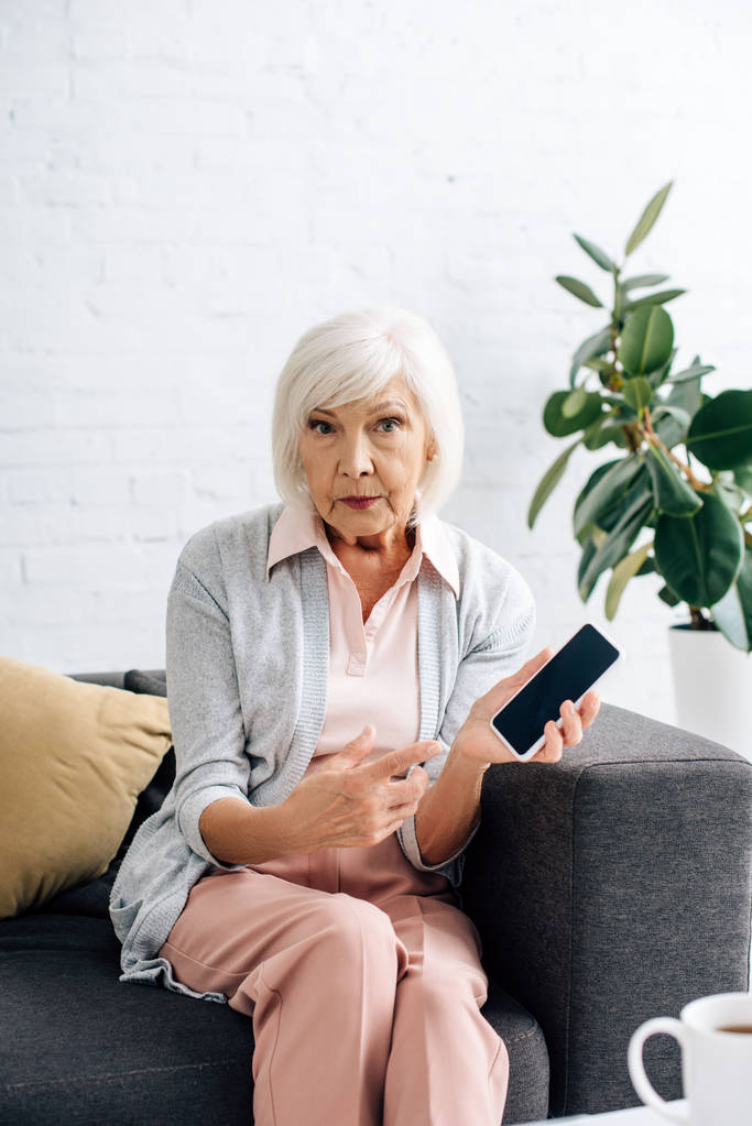 σοκαρισμένη ηλικιωμένη γυναίκα κάθεται στον καναπέ και δείχνοντας με το δάχτυλο στο smartphone στο διαμέρισμα  - Φωτογραφία, εικόνα