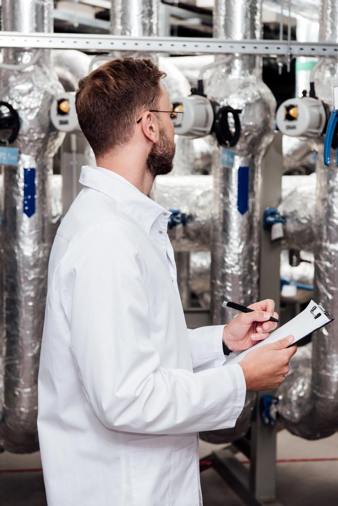bearded engineer in white coat holding clipboard and pen near air compressed system  - Photo, Image