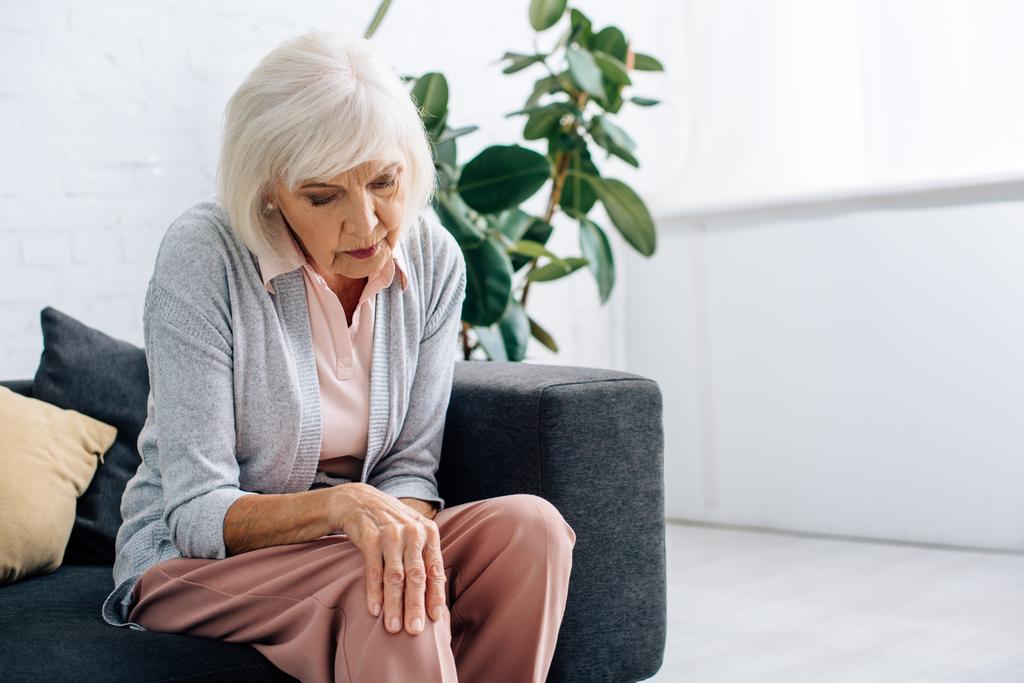 senior vrouw met knie Arthritis en zitten op de bank in appartement  - Foto, afbeelding