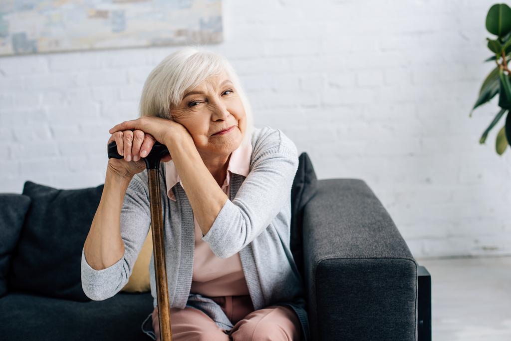 usmívající se starší žena s dřevěnou holí při pohledu na kameru v bytě  - Fotografie, Obrázek