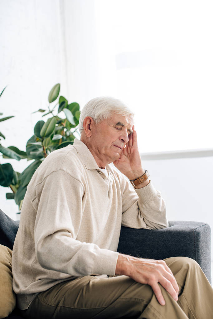 starszy mężczyzna siedzi na kanapie i ma ból głowy w mieszkaniu  - Zdjęcie, obraz