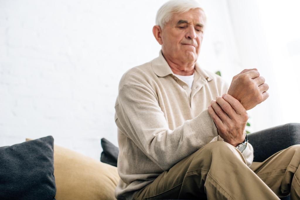 άποψη χαμηλή γωνία του ηλικιωμένου άνδρα που κάθεται στον καναπέ και έχοντας αρθρίτις στο χέρι στο διαμέρισμα  - Φωτογραφία, εικόνα