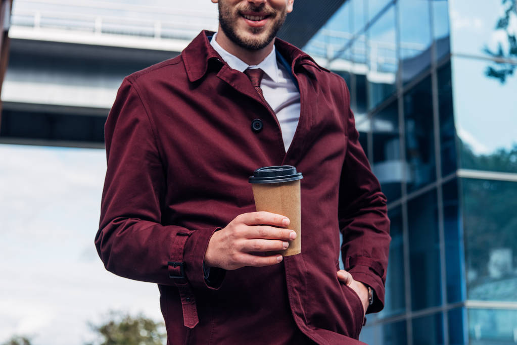 ostříhaný pohled na šťastný vousatý muž v kabátě drží papírový šálek, zatímco stojí s rukou v kapse  - Fotografie, Obrázek
