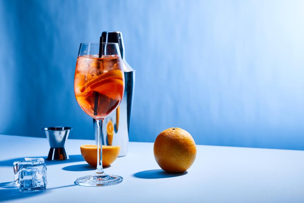 коктейль Aperol Spritz, апельсины, шейкер, кубики льда и измерительная чашка на синем фоне
  - Фото, изображение