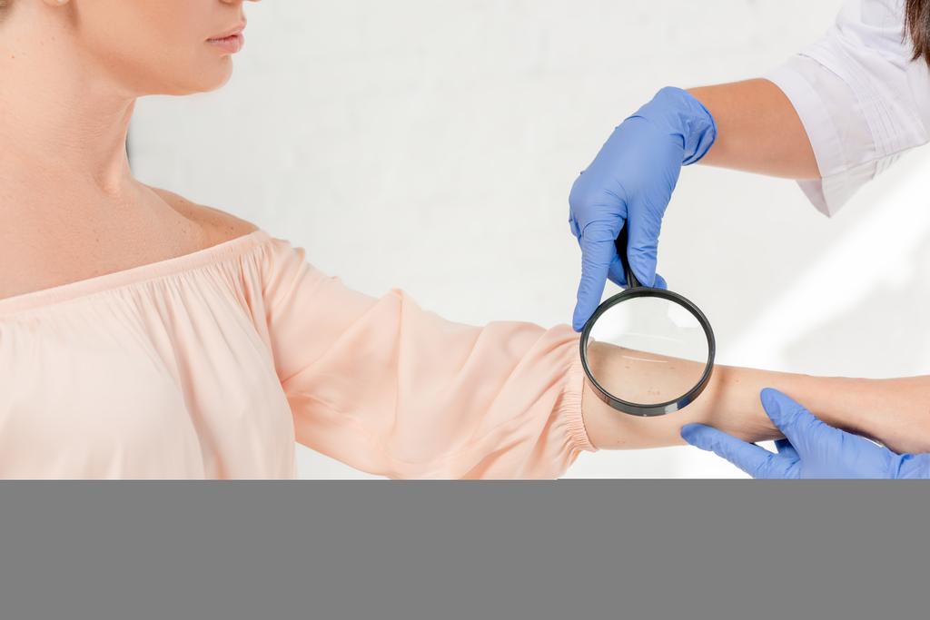 przycięty widok dermatologa badającego skórę pacjenta ze szkłem powiększającym w klinice - Zdjęcie, obraz