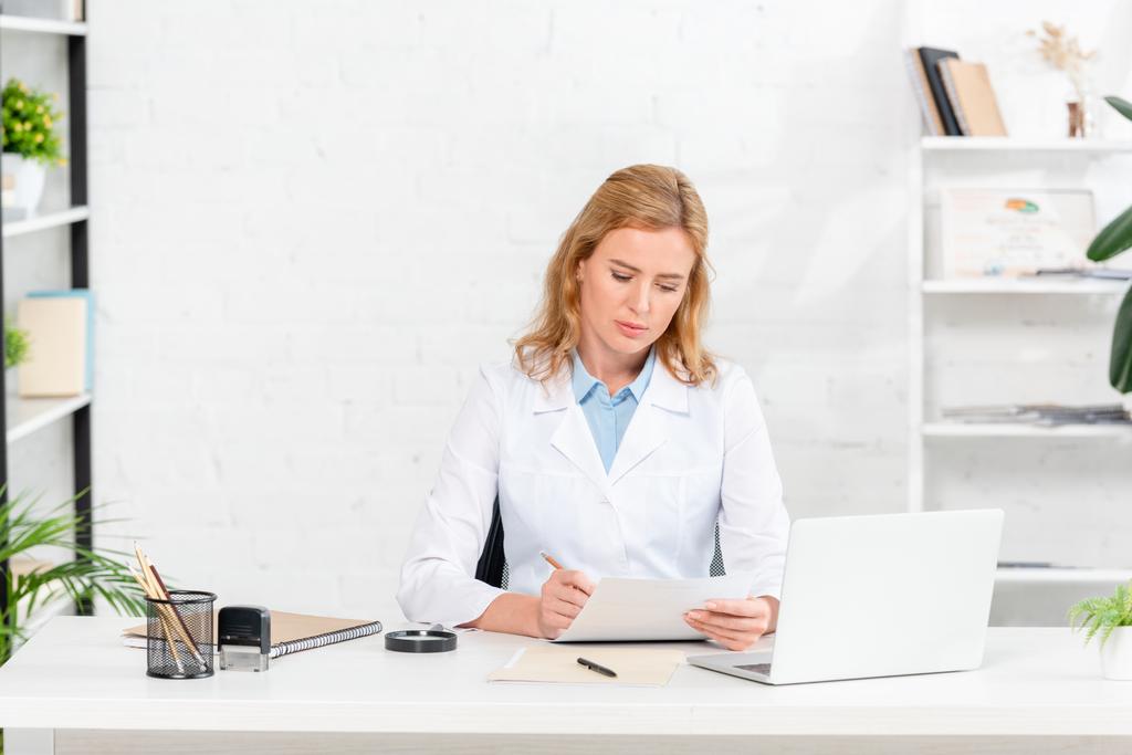 atractiva nutricionista sentada en la mesa y escribiendo en papel en la clínica
  - Foto, Imagen