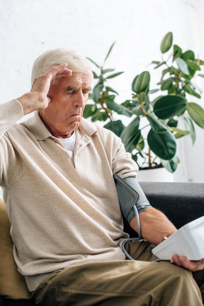 потрясенный пожилой человек измеряет артериальное давление и сидит на диване в квартире
  - Фото, изображение