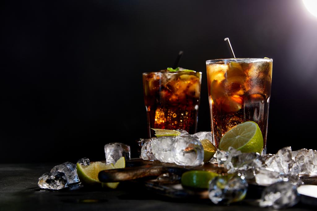 cocktails cuba libre σε ποτήρια με καλαμάκια, παγάκια και λάιμ σε μαύρο φόντο  - Φωτογραφία, εικόνα