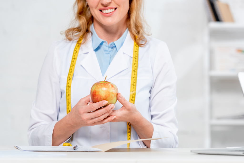 καλλιεργημένη άποψη του χαμογελαστού διατροφολόγου κάθεται στο τραπέζι και κρατώντας το μήλο στην κλινική  - Φωτογραφία, εικόνα