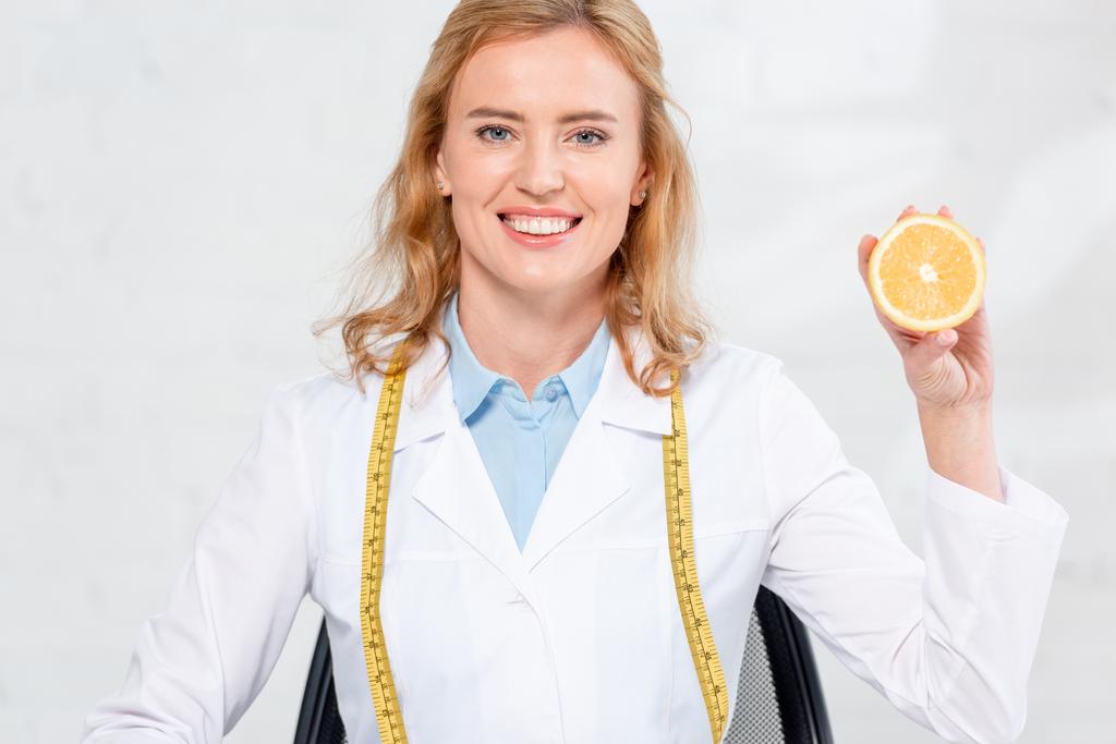 Улыбающийся диетолог с лентой, держащей срезанный апельсин в клинике
  - Фото, изображение