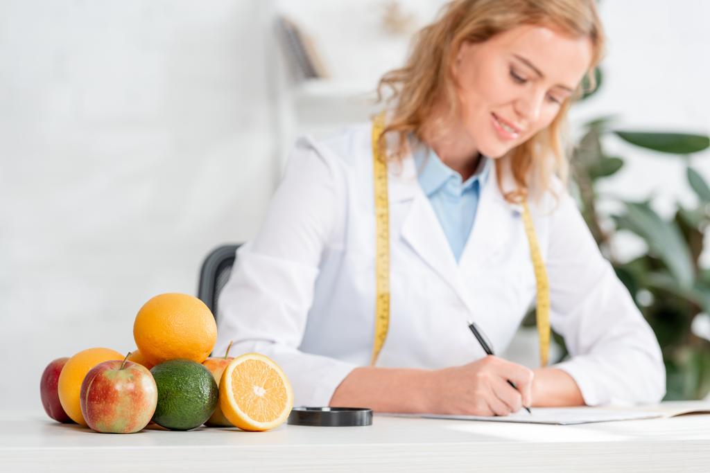 selektywne ukierunkowanie owoców i warzyw na stół i dietetyk siedzący przy stole i piszący na tle kliniki  - Zdjęcie, obraz