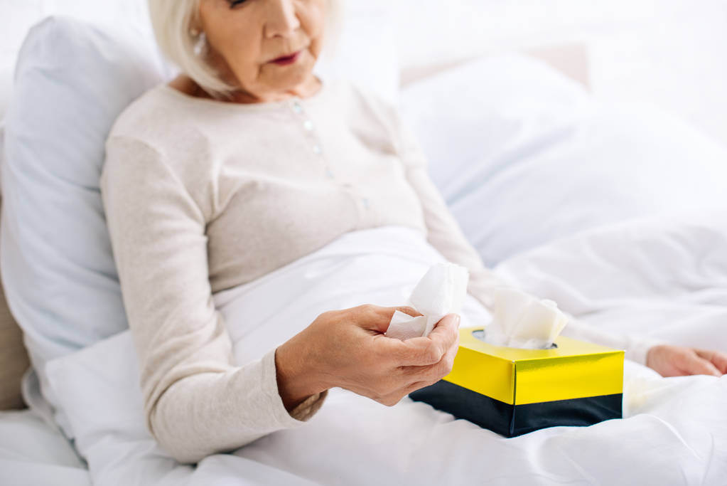 καλλιεργημένη άποψη της άρρωστης ηλικιωμένης γυναίκας που κρατά χαρτοπετσέτα στο κρεβάτι  - Φωτογραφία, εικόνα