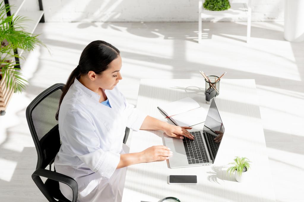 високий кут зору привабливого дерматолога, який сидить за столом і використовує ноутбук у клініці
  - Фото, зображення