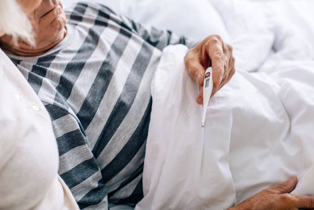 καλλιεργημένη άποψη του άρρωστου ηλικιωμένου άνδρα που κρατά θερμόμετρο στο κρεβάτι  - Φωτογραφία, εικόνα