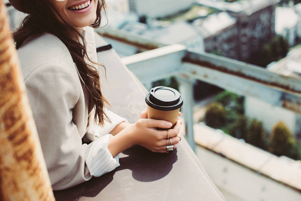 abgeschnittene Ansicht eines lächelnden modischen Mädchens im beigen Anzug, das auf dem Dach mit Coffee to go posiert - Foto, Bild