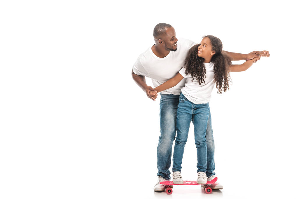 cheerful african american father supporting adorable daughter skateboarding on white background - Photo, Image