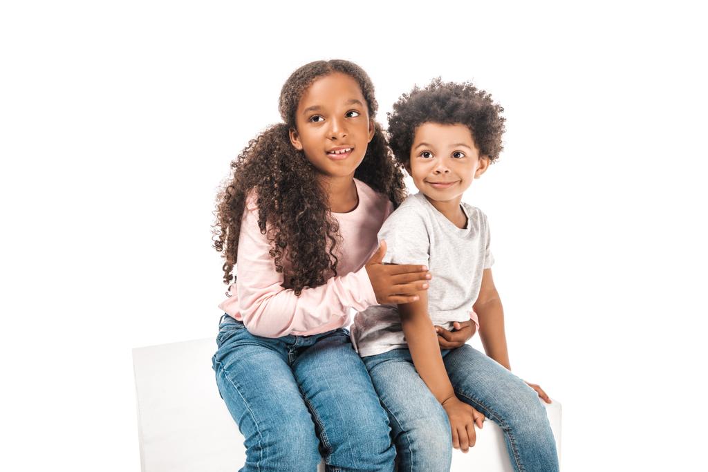 cute african american sister hugging adorable brother while sitting on pedestal and looking away together isolated on white - Photo, Image