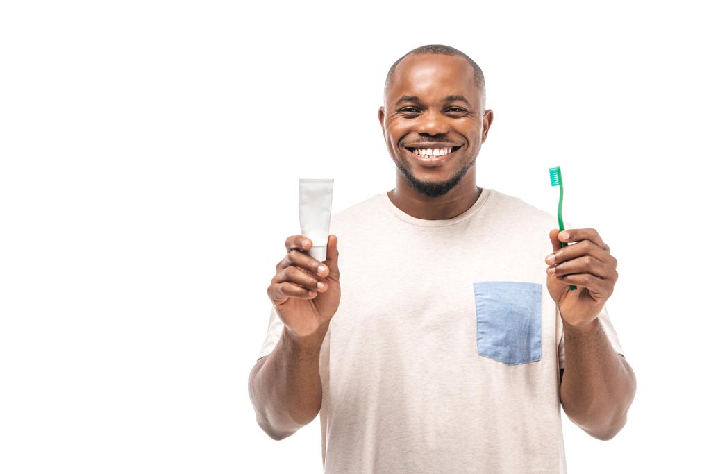 wesoły Afrykański Amerykanin trzymający szczoteczkę do zębów i pastę do zębów i patrzący na kamerę odizolowaną na biało - Zdjęcie, obraz