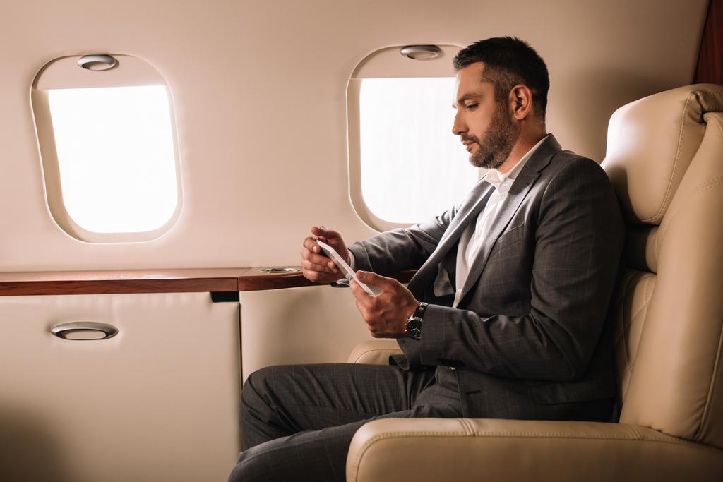 vista lateral del hombre de negocios guapo en traje usando teléfono inteligente en jet privado
  - Foto, imagen
