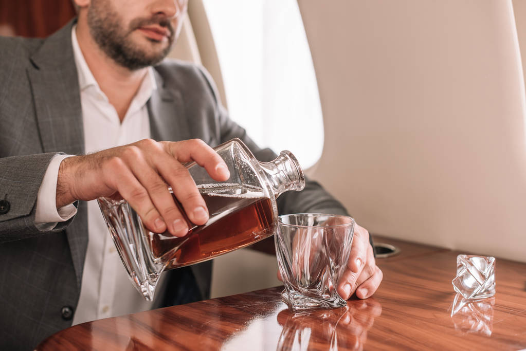 Ausgeschnittene Ansicht eines bärtigen Geschäftsmannes, der im Flugzeug Whiskey in Glas gießt  - Foto, Bild