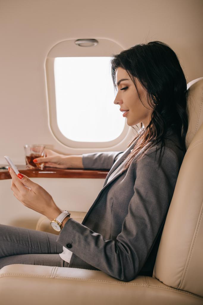 vista lateral de la mujer de negocios en el desgaste formal celebración de vidrio con whisky y teléfono inteligente en jet privado
  - Foto, Imagen