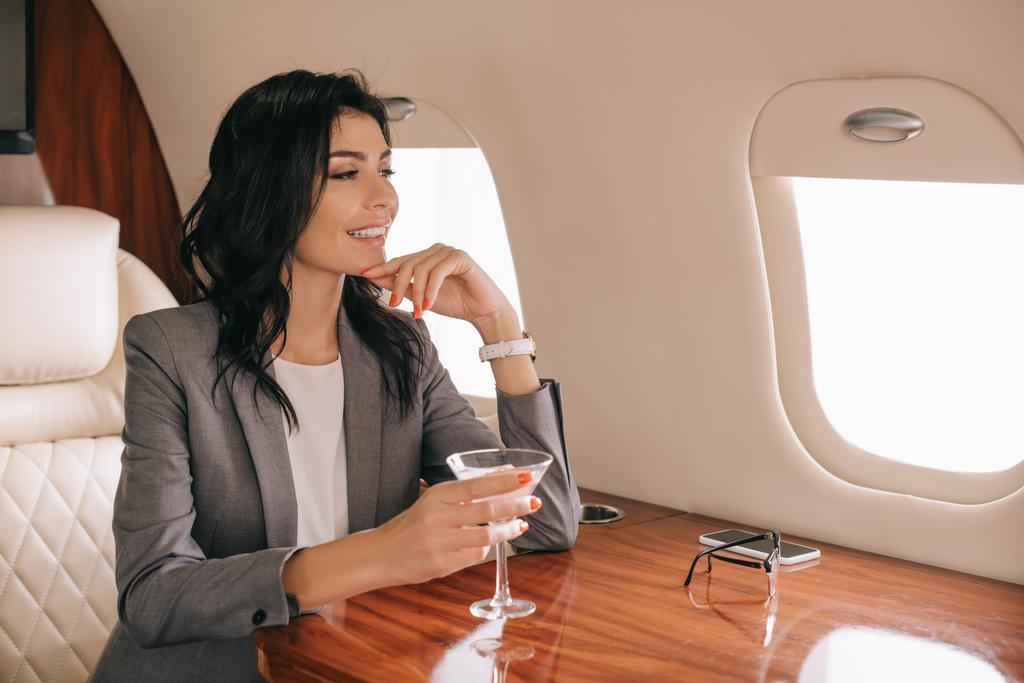 happy businesswoman in formal wear holding martini glass with alcohol in private jet  - 写真・画像