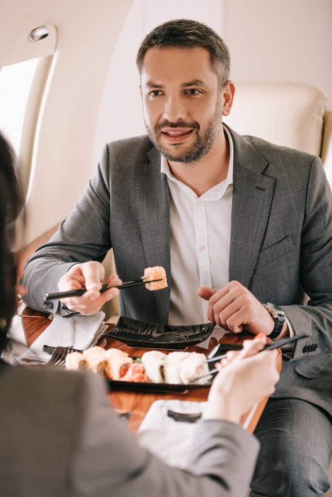 обрезанный вид бизнесвумен рядом с бородатым бизнесменом и вкусные суши в частном самолете
 - Фото, изображение