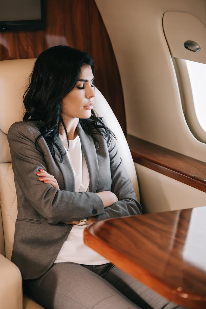 selective focus of attractive businesswoman with closed eyes and crossed arms sitting in private jet  - Photo, Image