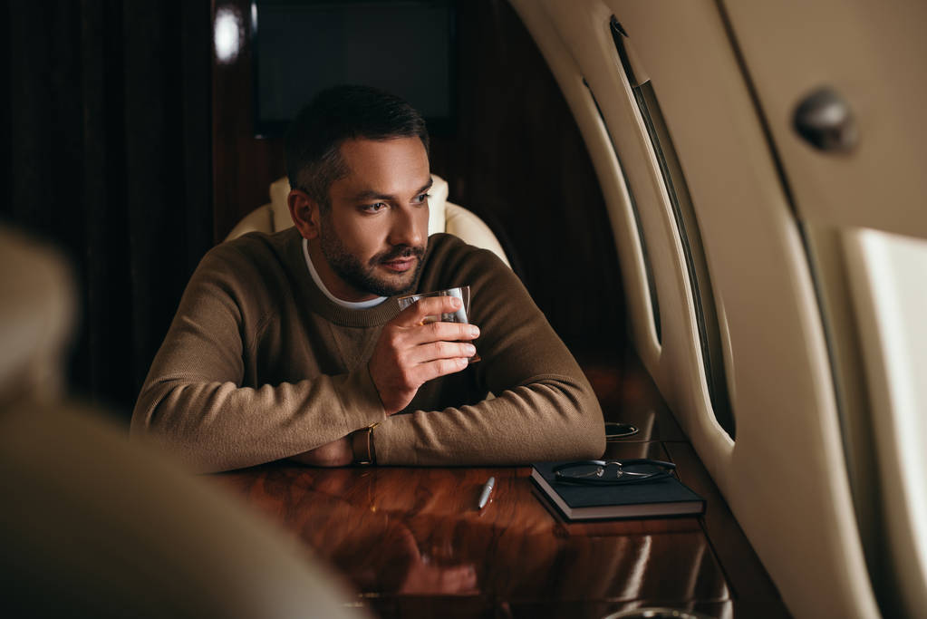 selective focus of handsome bearded man holding glass with alcohol drink while sitting in first class of private jet  - Photo, Image