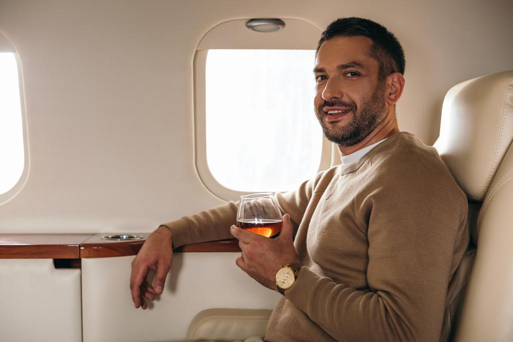 fröhlicher Mann hält Glas mit Cognac, während er in der ersten Klasse des Privatjets sitzt  - Foto, Bild