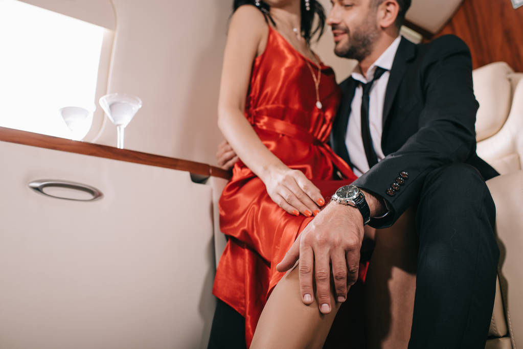περικοπή άποψη του γενειοφόρου άνδρα αγγίζοντας γυναίκα σε κόκκινο φόρεμα στην πρώτη θέση του ιδιωτικού τζετ  - Φωτογραφία, εικόνα